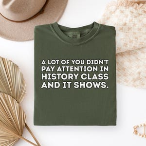 May include: An olive green sweatshirt with the text "A LOT OF YOU DIDN'T PAY ATTENTION IN HISTORY CLASS AND IT SHOWS." in white block letters. The sweatshirt is folded and rests on a cream-colored surface.