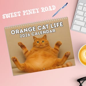 Op de afbeelding: Een spiraalgebonden kalender met de titel "ORANGE CAT LIFE 2026 CALENDAR" toont een oranje kat met uitgestrekte poten. De woorden "SWEET PINEY ROAD" staan bovenaan. Een pen, een toetsenbord en een kopje koffie zijn ook zichtbaar.