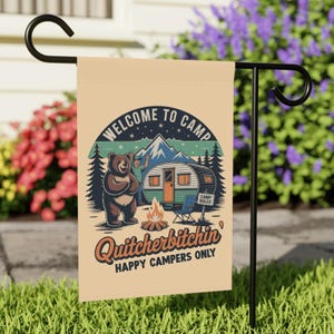 May include: A beige garden flag with a camping design. The flag shows a bear, a camper, a campfire, and the text "WELCOME TO CAMP" and "Quit yer bitchin' HAPPY CAMPERS ONLY". The flag is displayed on a black metal stand.