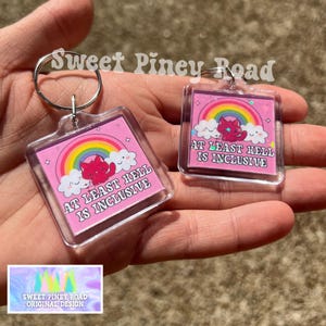 May include: Two square keychains with a pink background, rainbow, and a red devil illustration. The text "AT LEAST HELL IS INCLUSIVE" is displayed. The keychains are attached to silver keyrings. The brand name "Sweet Piney Road" is visible.