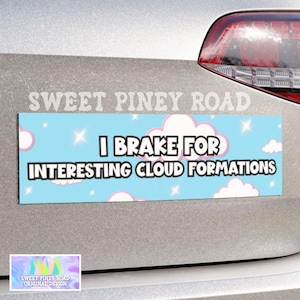 May include: A rectangular bumper sticker with a light blue background, white clouds, and stars. The text reads "I Brake for Interesting Cloud Formations" in black. The sticker is attached to a silver car.