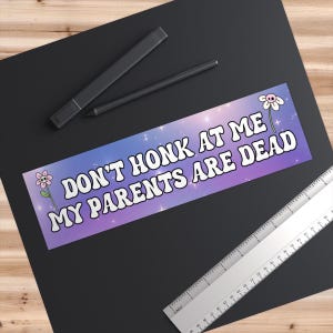 May include: A purple and blue bumper sticker with white stars and a white border. The sticker says "Don't honk at me my parents are dead" in white letters with a skull and crossbones flower graphic on each side.