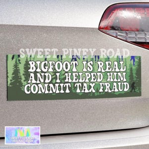 May include: A rectangular bumper sticker with the text "BIGFOOT IS REAL AND I HELPED HIM COMMIT TAX FRAUD" in white letters. The background is green with a forest design. The sticker is affixed to a silver vehicle.