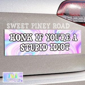 May include: A rectangular bumper sticker with the text "HONK IF YOU'RE A STUPID IDIOT" in bold white letters. The background has a swirling pattern of pink, purple, and blue. The words "SWEET PINEY ROAD" are above the sticker.