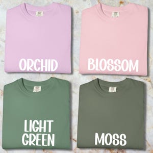 May include: Four t-shirts in shades of orchid, blossom, light green, and moss. Each has a crew neck and short sleeves. The colour names are printed in white block letters on each shirt.