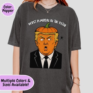 以下が含まれることがあります： ペッパーカラーのTシャツ。オレンジ色のカボチャの頭をした人物が黒いスーツとネクタイを着用しているグラフィックが描かれています。グラフィックの上部には「WORST PUMPKIN IN THE PATCH」という文字があり、近くにはクモの巣からぶら下がった小さな黒いクモが描かれています。