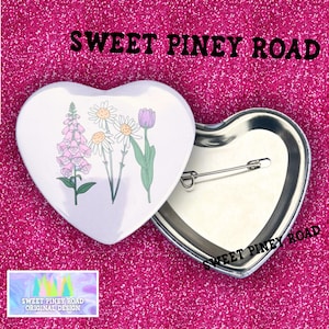 May include: Heart-shaped button with a floral design featuring pink foxgloves, white daisies, and a purple tulip. The text "SWEET PINEY ROAD" is above the button. The button is white with a metal pin back.