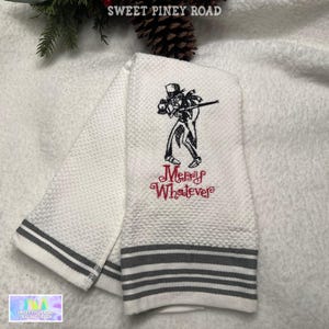 May include: White hand towel with a gray striped border and the embroidered phrase "Merry Whatever" in red. The towel features a black and white illustration of a skeleton in a top hat.