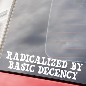 May include: White vinyl decal with the text "RADICALIZED BY BASIC DECENCY" in a bold, rounded font. The sticker is affixed to the rear window of a red vehicle, with the sky visible through the glass. The decal is designed to be applied to a car or other smooth surface.