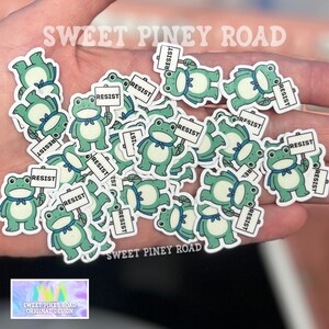 Puede incluir: Una colecci&oacute;n de pegatinas de ranas verdes, cada una con un letrero que dice "RESIST". Las ranas tienen pajaritas azules y est&aacute;n dispersas sobre una superficie. Tambi&eacute;n se ve el texto "SWEET PINEY ROAD" y "ORIGINAL DESIGN".