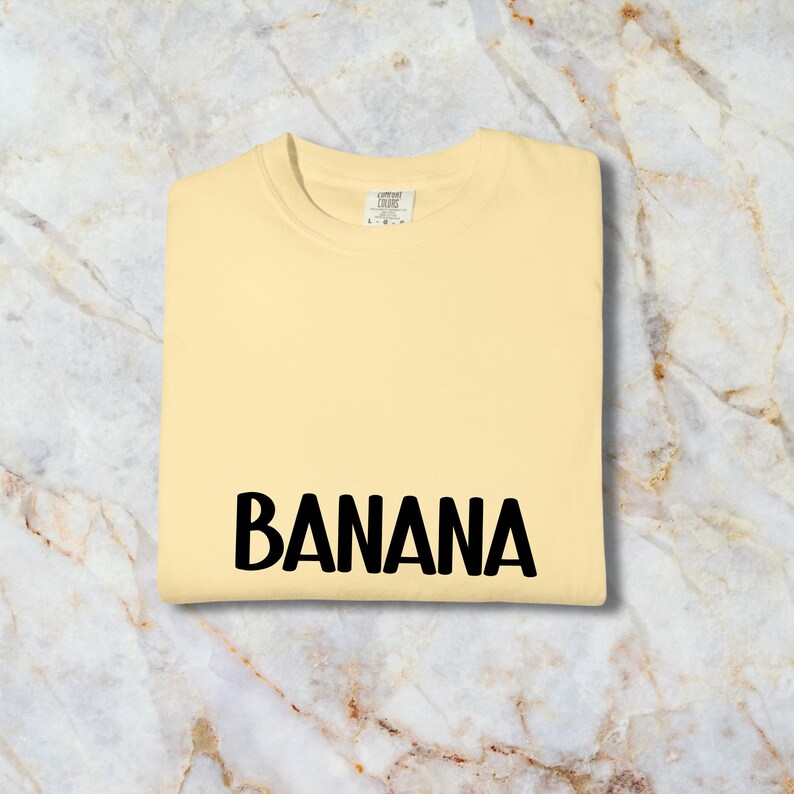 May include: A pale yellow t-shirt with the word "BANANA" printed in bold black letters. The shirt is folded neatly. The tag is visible in the upper centre of the shirt. The t-shirt appears to be made of a soft material.