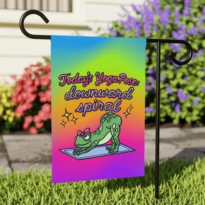 May include: A garden flag with a colorful gradient background and a cartoon frog in a yoga pose. The flag reads "Today's Yoga Pose: downward spiral" in a playful font. The frog is green with a pink bow.