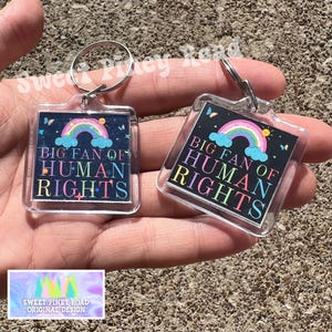 May include: Two clear acrylic keychains with silver key rings. Each keychain has a square design with a rainbow, clouds, and the text "BIG FAN OF HUMAN RIGHTS" in colorful letters. The background is dark blue.