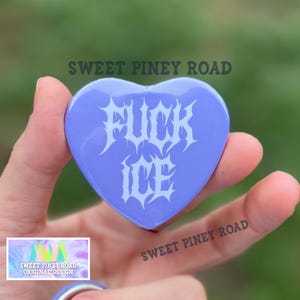 May include: A heart-shaped, light purple button with the words "FUCK ICE" in white, gothic-style lettering. The button is held in a hand, with the text "SWEET PINEY ROAD" visible. A smaller image with text is also visible.