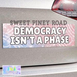 May include: A rectangular car bumper sticker with the text "SWEET PINEY ROAD DEMOCRACY ISN'T A PHASE" in white letters. The background has a pink, blue, and white glitter design. A small logo is in the bottom left corner.