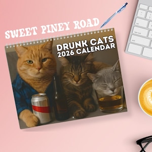 以下が含まれることがあります： 「DRUNK CATS 2026 CALENDAR」と書かれた、3匹の猫の写真が掲載されたリング綴じカレンダー。オレンジ色のトラ猫は青いシャツを着て缶を持ち、他の猫は茶色とグレー。背景はピンク色の表面。