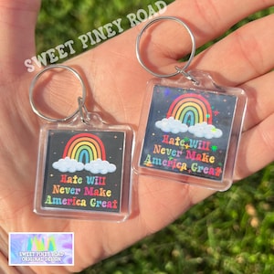 Peut inclure: Deux porte-clés carrés en acrylique transparent avec des anneaux argentés. Chaque porte-clés présente un arc-en-ciel au-dessus de nuages blancs sur fond noir, avec le texte "Hate Will Never Make America Great" en lettres colorées. Les porte-clés sont tenus dans une main.