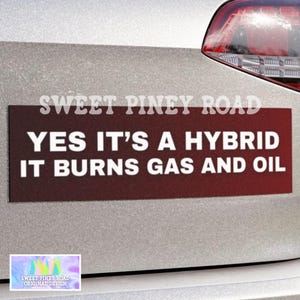 May include: A rectangular maroon bumper sticker with white text that reads "YES IT'S A HYBRID IT BURNS GAS AND OIL." The text "SWEET PINEY ROAD" is above the main text. A small sticker with the text "SWEET PINEY ROAD ORIGINAL DESIGN" is in the lower left corner.
