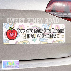 May include: A white car bumper sticker with a colorful design featuring a red apple, rainbows, and school supplies. The text reads "Teachers Make Tiny Humans Into Big Thinkers".