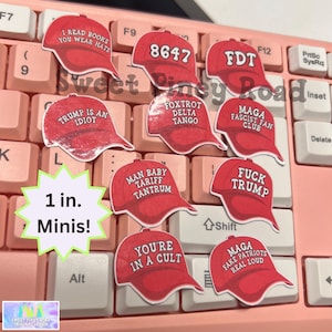 May include: A set of ten 1-inch red and white stickers featuring various political slogans and phrases. The stickers are shaped like baseball caps and are arranged on a pink keyboard.