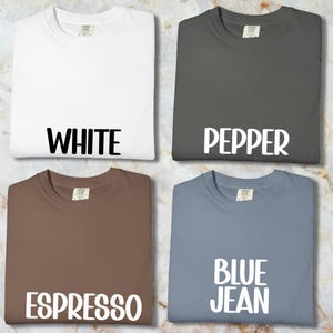 May include: Four folded t-shirts in white, pepper, espresso, and blue jean colours. Each shirt has the colour name printed in large, white, bold letters across the front. The shirts are displayed on a marble-patterned surface.