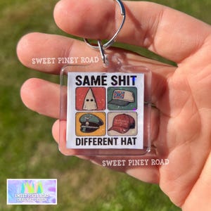 May include: Clear square keychain with a silver keyring. The keychain has the text "SAME SHIT DIFFERENT HAT" with four images of hats. The text "SWEET PINEY ROAD" is printed on the keychain.