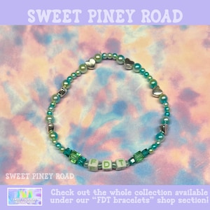 May include: A beaded bracelet with green, silver, and white beads. The bracelet features heart-shaped and square beads, with the letters "FDT" spelled out in white letter beads. The background is a tie-dye pattern with the text "SWEET PINEY ROAD".