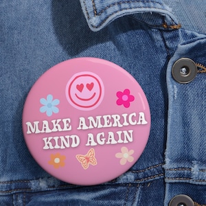 May include: A pink button with the text "MAKE AMERICA KIND AGAIN" in white. The button features a smiling face with heart eyes, along with flower and butterfly graphics in various colors. The button is set against a denim jacket.