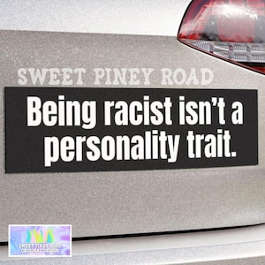 May include: A black rectangular bumper sticker with white text that reads, "Being racist isn't a personality trait." The sticker is affixed to a silver vehicle. The brand name "SWEET PINEY ROAD" is printed above the text.