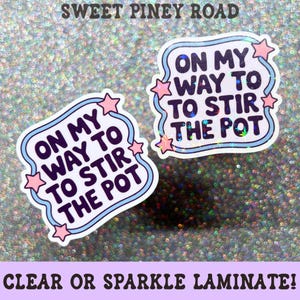 May include: Two die-cut stickers with a holographic finish featuring the text ON MY WAY TO STIR THE POT in dark purple letters. The stickers have a light blue wavy border and small pink stars at the corners, set against a glittery background.