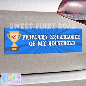 May include: A rectangular bumper sticker with a blue sunburst background. It features a gold trophy icon with a red no-bread symbol on the front. The white text reads: PRIMARY BREADLOSER OF MY HOUSEHOLD. The sticker is applied to a silver vehicle surface.