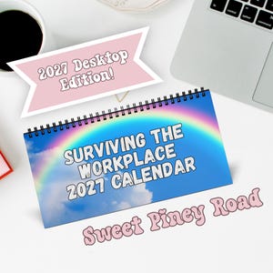 Op de afbeelding: Een spiraalgebonden bureaukalender getiteld Surviving the Workplace 2027 Calendar met een blauwe lucht en regenboog op de achtergrond. Een roze banner erboven leest 2027 Desktop Edition! en de tekst Sweet Piney Road staat in roze onder de kalender.