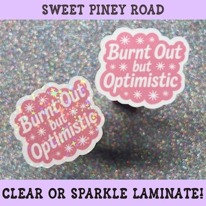 May include: Two scalloped-edge stickers featuring the white text Burnt Out but Optimistic on a pink background with white starburst accents. One sticker has a holographic sparkle finish, while the other has a smooth matte white finish.