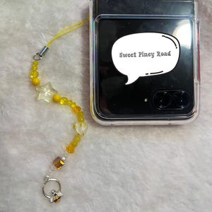 Puede incluir: Un charm de teléfono de cuentas amarillas y transparentes con un charm de abeja y una burbuja de diálogo que dice "Sweet Piney Road".