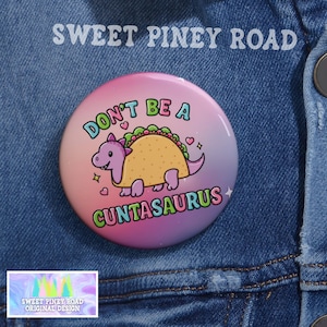 Puede incluir: Un botón redondo con un fondo degradado rosa, morado y amarillo. El botón presenta un dinosaurio de dibujos animados con una concha de taco, con el texto "DON'T BE A CUNTASAURUS". El texto "SWEET PINEY ROAD" está en la parte superior.