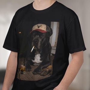 May include: Black t-shirt with a graphic of a black dog wearing a cap with a deer emblem and a cigarette. A glass of amber liquid is next to the dog. The dog has a white chest patch.