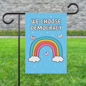 Könnte beinhalten: Eine blaue Gartenflagge mit einem Regenbogen und Schmetterlingen. Der Text auf der Flagge lautet "We Choose Democracy".