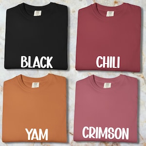 May include: Four plain t-shirts in black, chili, yam, and crimson. Each shirt has a crew neck and the colour name printed in white block letters. The shirts are displayed on a marble-patterned surface.