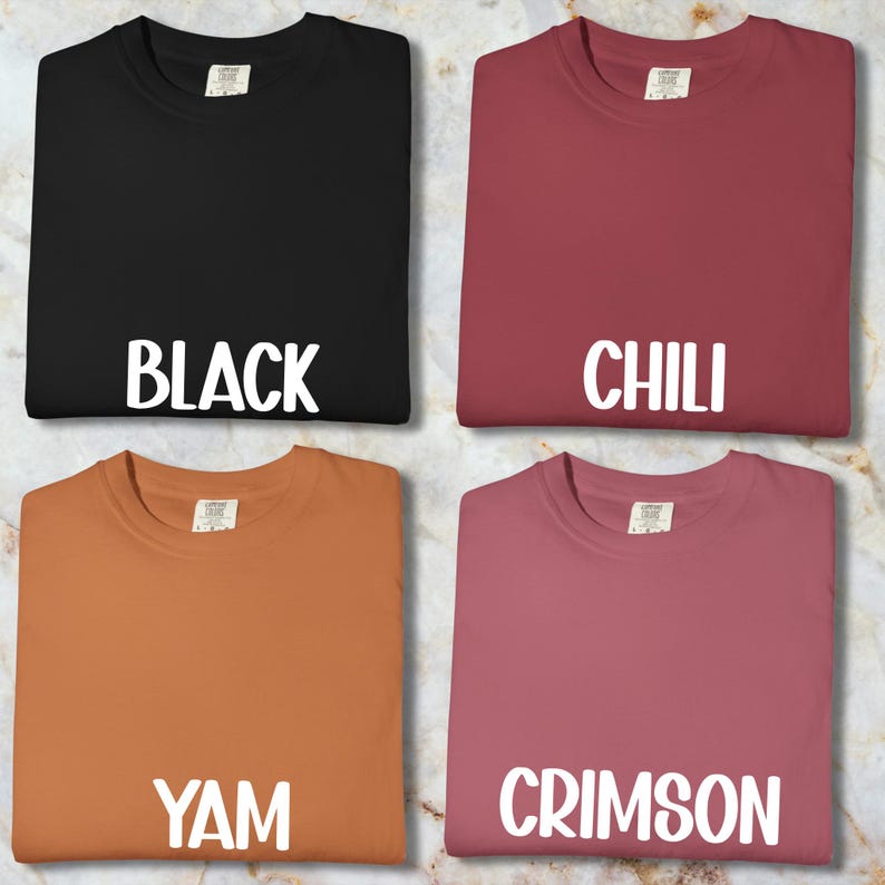 May include: Four plain t-shirts in black, chili, yam, and crimson. Each shirt has a crew neck and the colour name printed in white block letters. The shirts are displayed on a marble-patterned surface.