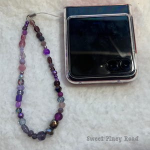 Op de afbeelding: Een paarse en zilveren kralen telefoonbedel met een transparante hoes voor een opvouwbare telefoon. De telefoonhoes heeft een zwart scherm en twee camera lenzen. De tekst "Sweet Piney Road" is zichtbaar in de rechter benedenhoek van de afbeelding.