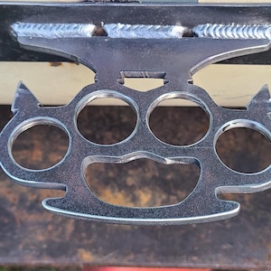 Op de afbeelding: Een zilverkleurige metalen boksbeugel met vier vingergaten en een gestileerd ontwerp. De knokkels zijn bevestigd aan een montageplaat met gelaste naden. Het object wordt tegen een donkere achtergrond getoond.