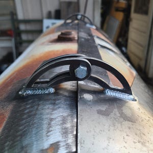 Puede incluir: Primer plano de un ahumador de barbacoa de metal con acabado oxidado. El ahumador presenta un asa curva asegurada con un perno. El metal tiene una mezcla de tonos plateados y marrones, con marcas de soldadura visibles. Diseño artesanal.