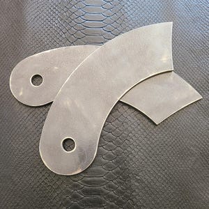 Pode incluir: Duas peças de metal com bordas curvas e um furo no centro. As peças são de cor cinza claro e têm uma textura áspera.