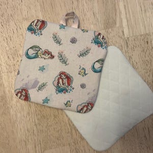 May include: A square, light-colored pot holder with a loop handle, featuring a repeating pattern of a mermaid, sea creatures, and aquatic plants. A matching, quilted, triangular pot holder is next to it.