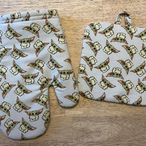 May include: Oven mitt and pot holder set featuring a repeating pattern of a cartoon character with large ears and eyes on a light gray background. The oven mitt has a loop for hanging.