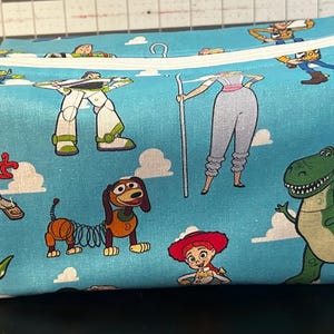 Puede incluir: Una bolsa rectangular de tela azul claro con personajes de dibujos animados de la franquicia Toy Story. La bolsa tiene una cremallera blanca y un peque&ntilde;o lazo. Los personajes incluyen a Woody, Buzz Lightyear, Rex y otros, sobre un fondo de cielo y nubes.