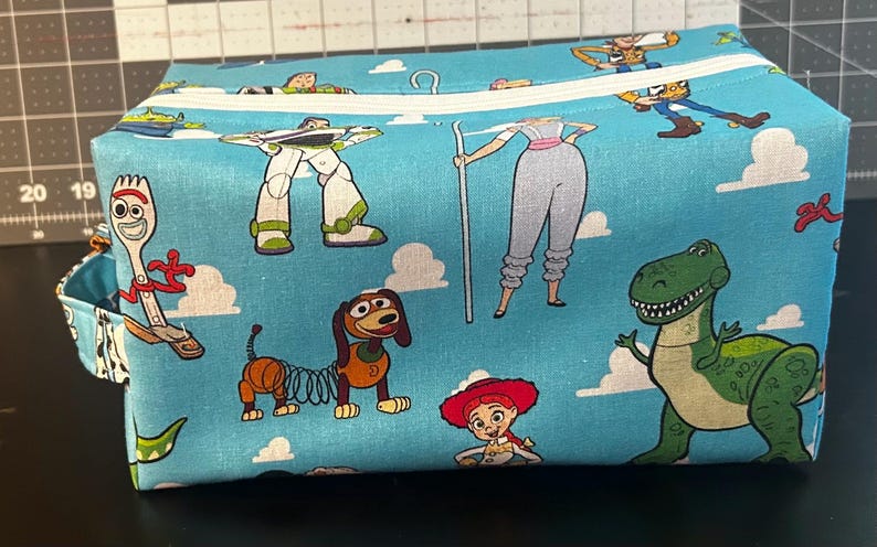 Puede incluir: Una bolsa rectangular de tela azul claro con personajes de dibujos animados de la franquicia Toy Story. La bolsa tiene una cremallera blanca y un peque&ntilde;o lazo. Los personajes incluyen a Woody, Buzz Lightyear, Rex y otros, sobre un fondo de cielo y nubes.