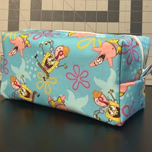 May include: A rectangular, turquoise fabric cosmetic bag featuring a repeating pattern of SpongeBob SquarePants, Patrick Star, and floral designs. The bag has a white zipper and a small fabric loop on the side.