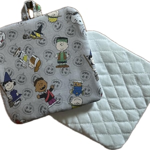 May include: A gray, square pot holder with a loop, featuring a Halloween-themed print of cartoon characters and jack-o'-lanterns. A second, light gray, quilted pot holder is visible next to it. The pot holder is approximately 8 inches square.