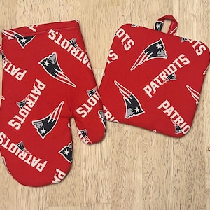 May include: A red oven mitt and pot holder set. The set features a repeating pattern of the word "PATRIOTS" in white, along with the team logo. The oven mitt has a loop for hanging.